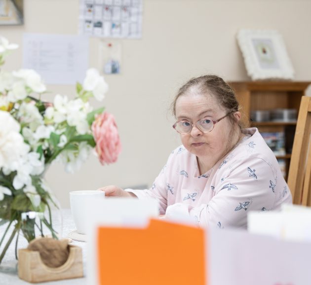 Lady sitting at a table with flowers. She is looking at the camera.