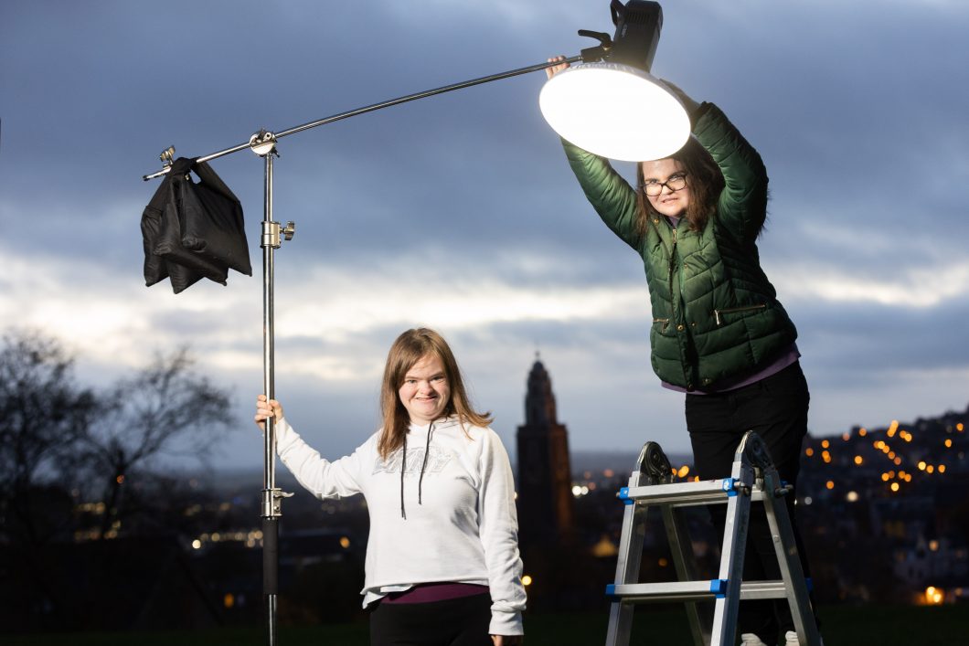 Two young woman outside. It is dusk. The woman on the left is holding a light and the woman on the right has turned on the light., she is standing on a ladder to reach it.