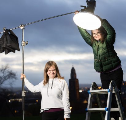 Two young woman outside. It is dusk. The woman on the left is holding a light and the woman on the right has turned on the light., she is standing on a ladder to reach it.